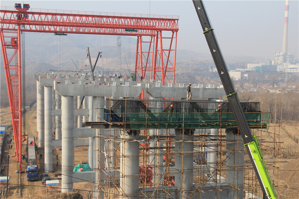 京新高速公路南洋河特大桥施工现场 本次架设T梁长40米,重达156吨,位于南洋河特大桥8号——9号墩之间。南洋特大桥桥长1073米,沿线路走向依次上跨南洋河、S247省道、旧S247省道及京包铁路。主桥为整幅设计,采用现浇混凝土转体箱梁结构,转体跨径2*55米,转体吨位重达15000吨。桥面为双向六车道。引桥为分幅设计,采用40米及35米 T梁,单幅单孔7片,共336片,利用提梁门吊及架桥机配合进行T梁架设。南洋河特大桥为京新高速公路的重要组成部分,也是全线唯一一座跨京包铁路转体